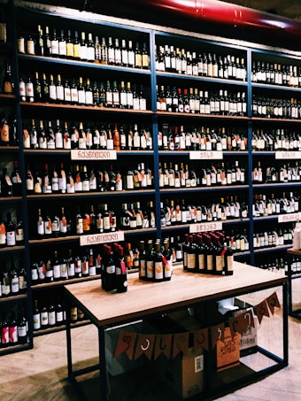 Shelves filled with various bottles of wine displayed in a store. The labels on the bottles and the signs above the shelves suggest a diverse selection. A table in the foreground exhibits additional bottles, and boxes are stacked below it with banners hanging down.
