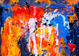 A vibrant abstract painting featuring bold strokes and splatters of color. Predominant hues include orange, red, and blue, with intertwined black lines adding movement and contrast. The texture is dynamic and expressive, showcasing layers of paint applied with energy and spontaneity.