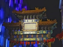 An ornate, traditional Chinese archway is prominently displayed against a dark backdrop. The structure is richly decorated with intricate patterns, vibrant colors, and detailed ornamentation. Hanging red lanterns are visible beneath the archway, adding to its elaborate appearance. The background features illuminated vertical lines in shades of blue and purple, enhancing the visual contrast.