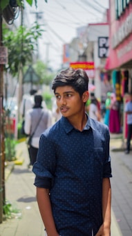 A young person stands on a bustling street, wearing a dark blue shirt with rolled-up sleeves. The background is lively with people walking, colorful signs, and shops lining the street. There is greenery and electrical wires visible as well.