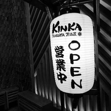 A large, cylindrical paper lantern illuminated, displaying the text 'KINKA IZAKAYA OPEN' in English, alongside Japanese characters. The lantern is hung against a background of textured walls with wooden paneling.