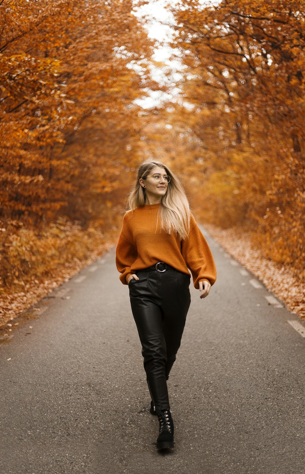 woman in orange sweater and black pants walking on street between orange trees