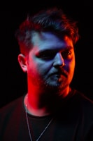 A portrait of a man with a beard is lit with dramatic lighting. The face is illuminated by red and blue lights, creating a striking contrast on the skin. The background is completely black, enhancing the vividness of the colors on the subject's face.