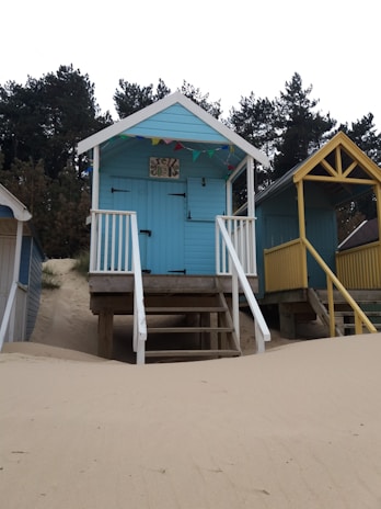 A row of colorful beach huts elevated on stilts, surrounded by sandy dunes and pine trees. The huts feature decorated entrances with small staircases and bunting overhead, creating a seaside atmosphere.