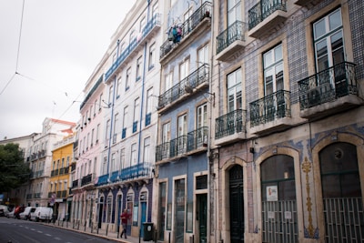 A row of colorful, historic buildings with wrought iron balconies lines a street. The architecture features intricate tile work and a variety of pastel colors including blue, pink, and yellow. Cars and people are visible on the street, with a tree in the background adding greenery to the urban scene.