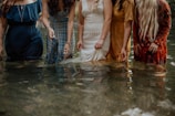 Five women stand in knee-deep water, each wearing a different dress. The dresses are of various textures and colors, including blue, plaid, white lace, yellow, and red with patterns. They are partially submerged, creating a reflective surface on the water.