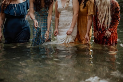 Five women stand in knee-deep water, each wearing a different dress. The dresses are of various textures and colors, including blue, plaid, white lace, yellow, and red with patterns. They are partially submerged, creating a reflective surface on the water.