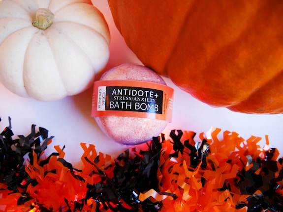 A pink colored bath bomb labeled 'Antidote Stress/Anxiety Bath Bomb' with a 'Pumpkin Spice' tag is placed between a white and an orange pumpkin. Surrounding these items is decorative shredded paper in black and orange colors, creating a festive atmosphere.