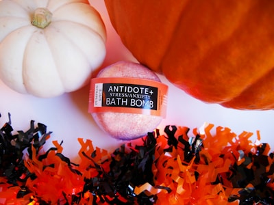 A pink colored bath bomb labeled 'Antidote Stress/Anxiety Bath Bomb' with a 'Pumpkin Spice' tag is placed between a white and an orange pumpkin. Surrounding these items is decorative shredded paper in black and orange colors, creating a festive atmosphere.