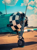 A vintage propeller airplane with a checkered pattern on the nose is parked on a sunlit airstrip. The airplane has a teal color, and the background features a bright blue sky with scattered white clouds.