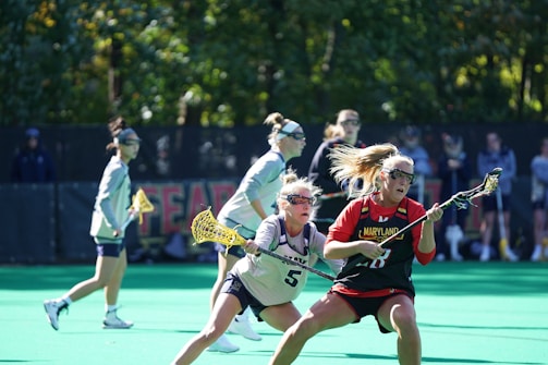A group of female lacrosse players are actively engaged in a game on a field. One player in a Maryland jersey is in possession of the ball, while another player in a helmet is closely defending her. Additional players are visible in the background, focused on the action. The field is green, and trees are seen in the background, providing a natural setting.