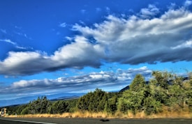 The image depicts a scenic landscape with a clear, vibrant blue sky filled with scattered, fluffy white clouds. Below the sky, there is an expansive view of green trees and bushes alongside a road that runs parallel to the greenery. In the background, there are distant mountains visible on the horizon.