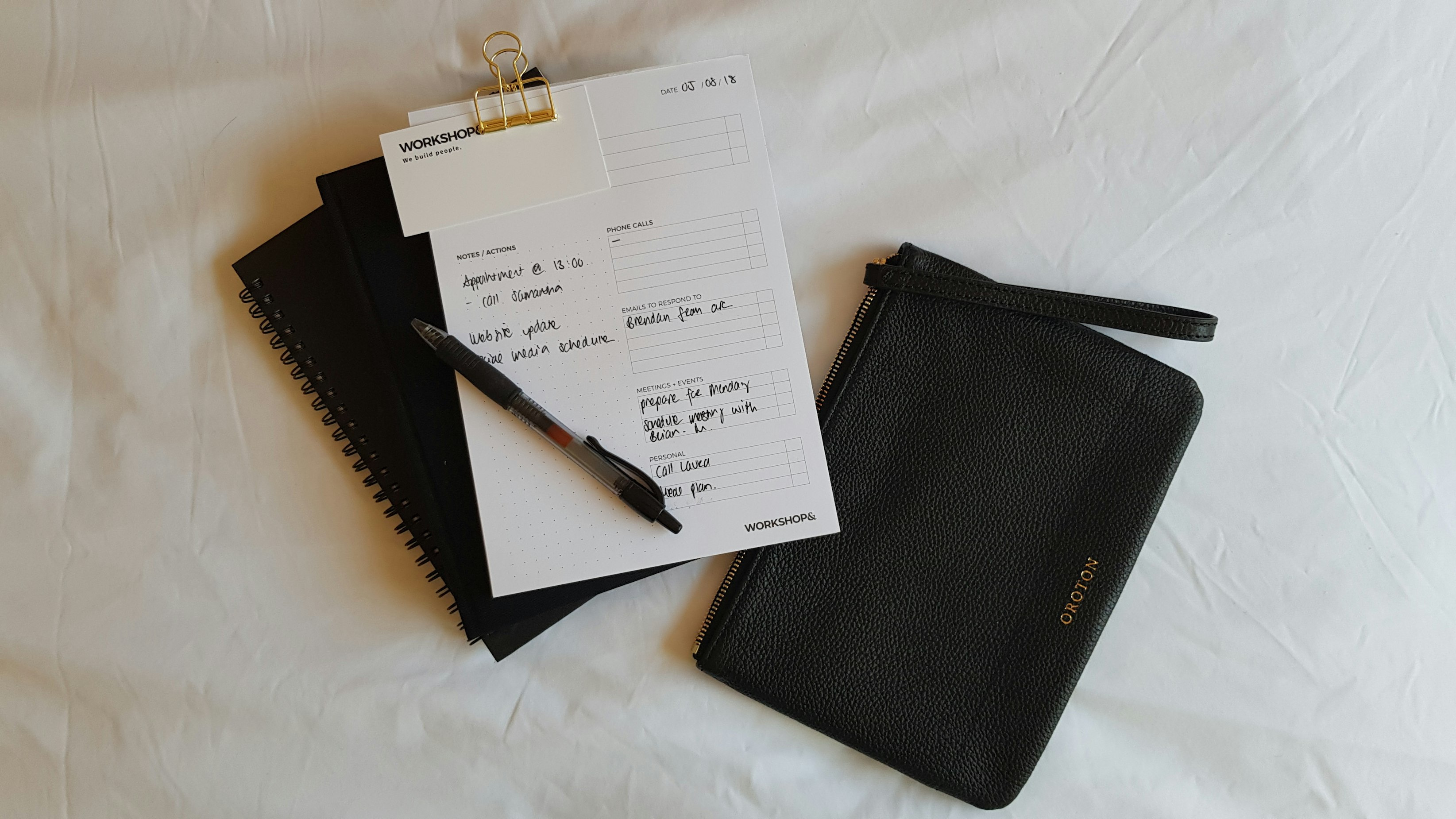 A note book with writing on it, on a white bed, with a small black purse next to it.