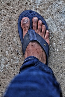 A dirty foot in a worn-out blue flip-flop is visible, set against a rough concrete ground. The skin appears rough and slightly dirty, with visible markings or scuffs. The jeans visible in the image are a dark blue denim.