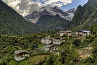 Nestled in a lush green valley, a small village is surrounded by dense vegetation and towering mountains. Traditional houses with sloped roofs are scattered across the terrain, indicating a rural and serene lifestyle. In the background, majestic snow-capped peaks rise above the scene, partially shrouded in clouds. The overall atmosphere is tranquil and idyllic.