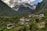 Nestled in a lush green valley, a small village is surrounded by dense vegetation and towering mountains. Traditional houses with sloped roofs are scattered across the terrain, indicating a rural and serene lifestyle. In the background, majestic snow-capped peaks rise above the scene, partially shrouded in clouds. The overall atmosphere is tranquil and idyllic.