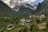 Nestled in a lush green valley, a small village is surrounded by dense vegetation and towering mountains. Traditional houses with sloped roofs are scattered across the terrain, indicating a rural and serene lifestyle. In the background, majestic snow-capped peaks rise above the scene, partially shrouded in clouds. The overall atmosphere is tranquil and idyllic.