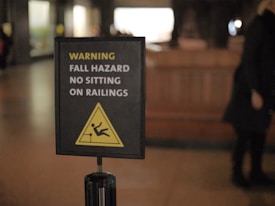 A warning sign indicating a fall hazard with instructions not to sit on railings. The sign displays a yellow warning symbol with a figure slipping off a railing.