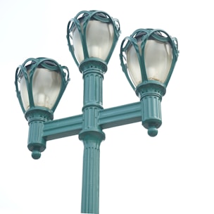 A vintage-style street lamp with three ornate globes is mounted on a tall, textured pole. The lamps have a blue-green color with decorative metalwork encasing the frosted glass of the globes.