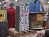 A vintage postage stamp dispenser is displayed on a wooden table surrounded by other antique items. The dispenser is labeled 'Postage Stamps' and has slots for different denominations. There are boxes and signs with retro designs in the background, and a person in a red coat stands blurred in the background.