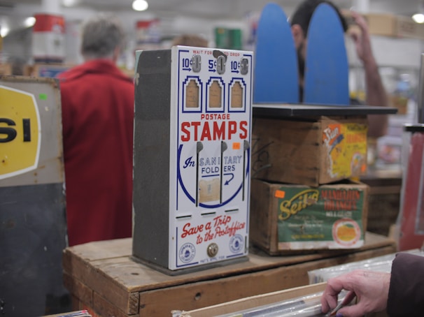 A vintage postage stamp dispenser is displayed on a wooden table surrounded by other antique items. The dispenser is labeled 'Postage Stamps' and has slots for different denominations. There are boxes and signs with retro designs in the background, and a person in a red coat stands blurred in the background.