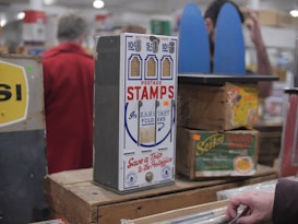 A vintage postage stamp dispenser is displayed on a wooden table surrounded by other antique items. The dispenser is labeled 'Postage Stamps' and has slots for different denominations. There are boxes and signs with retro designs in the background, and a person in a red coat stands blurred in the background.