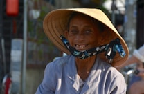 An elderly individual with a warm and genuine smile wears a traditional straw hat and a light blue shirt. The background is slightly blurred, with hints of greenery and light creating a serene setting. The person’s face is marked with wrinkles, suggesting wisdom and experience.