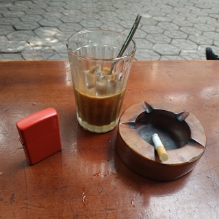 On a wooden table, there is a glass of coffee with a metal spoon inside, a red lighter, and a wooden ashtray with a cigarette. The ground is paved with stone tiles.
