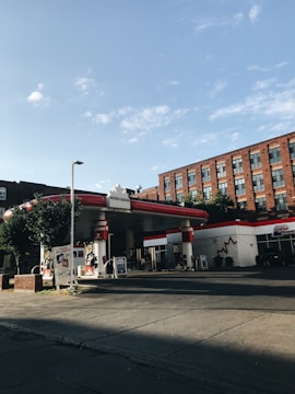 A gas station with a distinctive red and white color scheme is positioned on a street corner, adjacent to a brick building with multiple windows. Trees and a clear blue sky add to the scene's calm atmosphere.