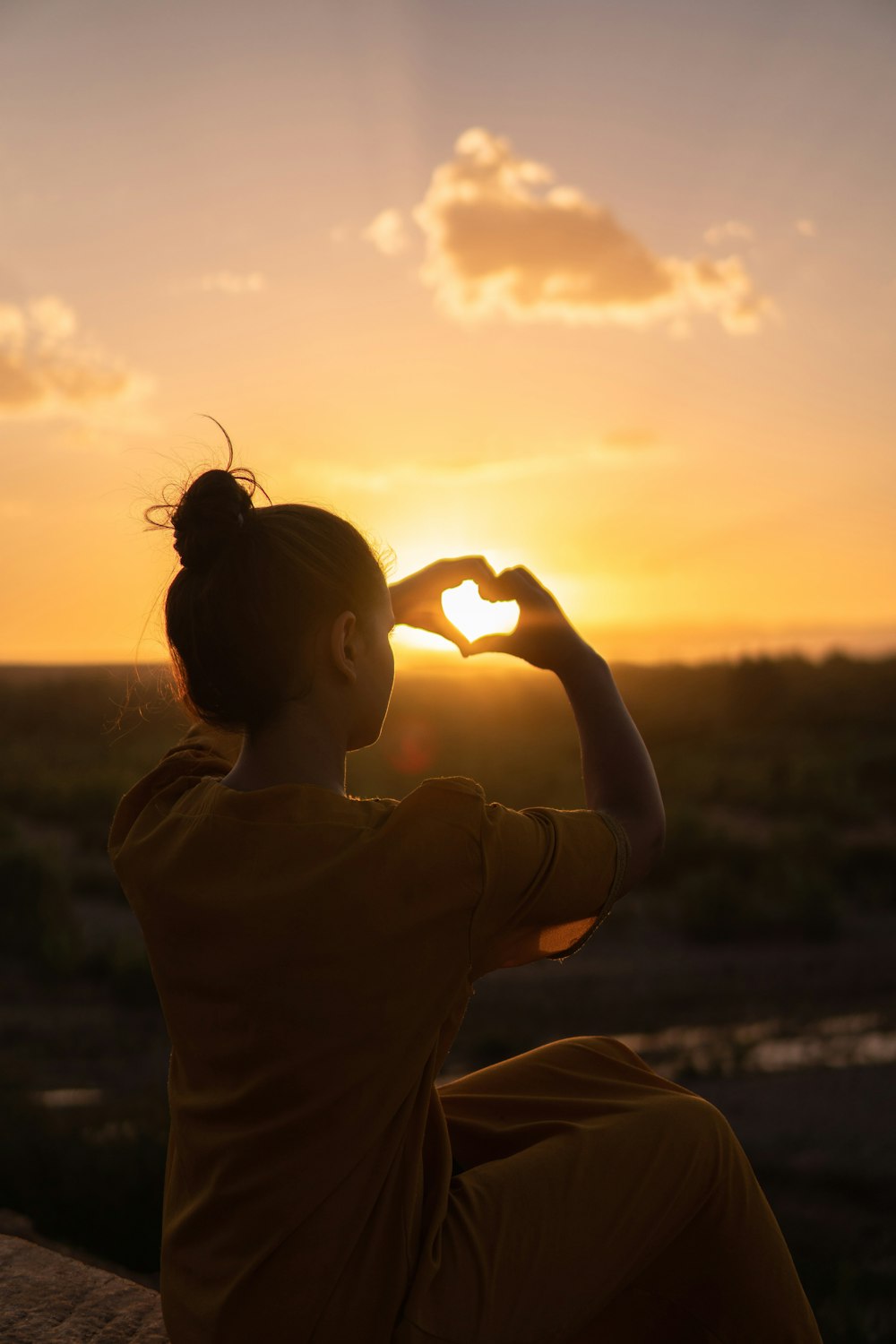 woman sitting at the cliff edge forming heart hand sign on sunset sun