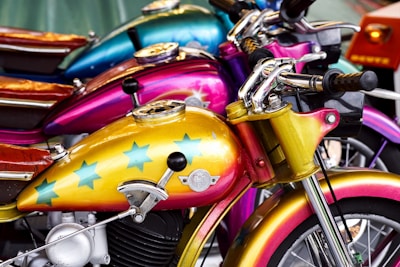 A row of vibrantly colored motorcycles lined up next to each other. The foremost motorcycle features a golden-yellow tank adorned with blue stars and shiny chrome details. Behind it, other bikes display shades of pink and blue with similar chrome accents. The seats are made of rich, brown leather.