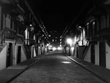 A quiet, empty street lined with symmetrical rows of old brownstone buildings. The scene is captured in black and white, highlighting the contrast between the bright streetlights and the shadowy facades. The cobblestone road extends into the distance, with stairs leading up to the entrances of each building.