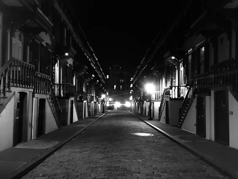 A quiet, empty street lined with symmetrical rows of old brownstone buildings. The scene is captured in black and white, highlighting the contrast between the bright streetlights and the shadowy facades. The cobblestone road extends into the distance, with stairs leading up to the entrances of each building.