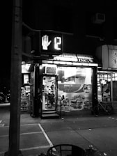 A nighttime street scene with a small deli called 'Amsterdam Deli'. The deli has bright signage, and a street crossing signal showing a hand and the number 12 is visible. There is a bicycle parked outside and various advertisements on the deli windows.