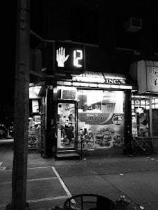 A nighttime street scene with a small deli called 'Amsterdam Deli'. The deli has bright signage, and a street crossing signal showing a hand and the number 12 is visible. There is a bicycle parked outside and various advertisements on the deli windows.