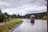 A scenic view of a tranquil reservoir surrounded by lush green forest and a cloudy sky. A pathway lined with grass and rocks runs alongside the water, where people are jogging and walking a dog. A small brick building with a pointed roof is situated on the water, accessible by a narrow bridge. The scene conveys a peaceful, natural setting with an emphasis on outdoor recreation.