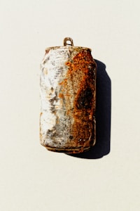 A highly rusted and weathered aluminum can with visible oxidation, placed against a plain background with a shadow cast to one side.