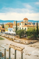 A scenic view of a Mediterranean landscape featuring traditional architecture. In the foreground, there are ancient stone ruins and columns. The background showcases colorful buildings with red-tiled roofs, surrounded by trees and greenery. The sky is partly cloudy, adding to the serene atmosphere.
