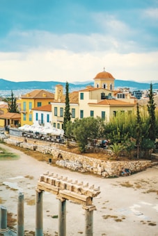 A scenic view of a Mediterranean landscape featuring traditional architecture. In the foreground, there are ancient stone ruins and columns. The background showcases colorful buildings with red-tiled roofs, surrounded by trees and greenery. The sky is partly cloudy, adding to the serene atmosphere.