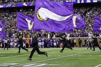 A group of individuals dressed in black are running across a football field, holding large, purple flags. The flags feature designs, including a white horn, associated with a sports team. In the background, a large crowd of spectators dressed in purple and yellow fills the stadium stands.