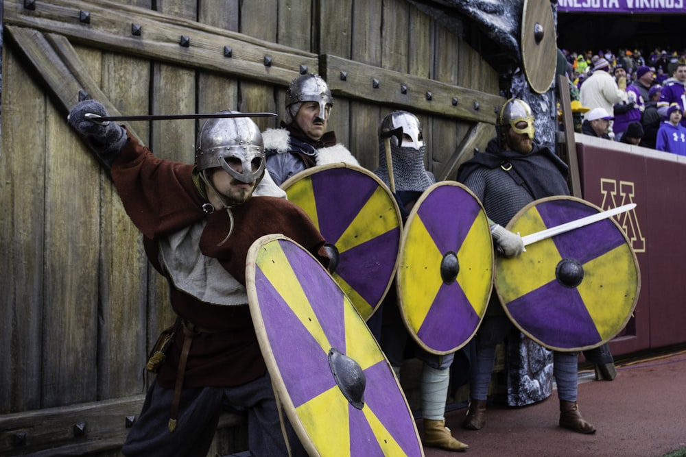 four men wearing armor holding sword and shield standing in front of wall