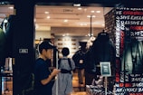 A person wearing a cap and backpack is looking at their phone while walking past a clothing store. The store has a display of jackets and coats, and the interior is well-lit with warm tones. The word 'EXCLUSIVE' is visible on the wall, and there is a black puffer jacket prominently displayed.