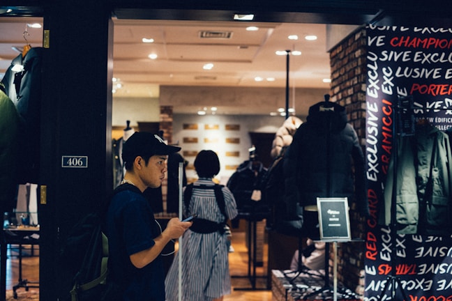 A person wearing a cap and backpack is looking at their phone while walking past a clothing store. The store has a display of jackets and coats, and the interior is well-lit with warm tones. The word 'EXCLUSIVE' is visible on the wall, and there is a black puffer jacket prominently displayed.