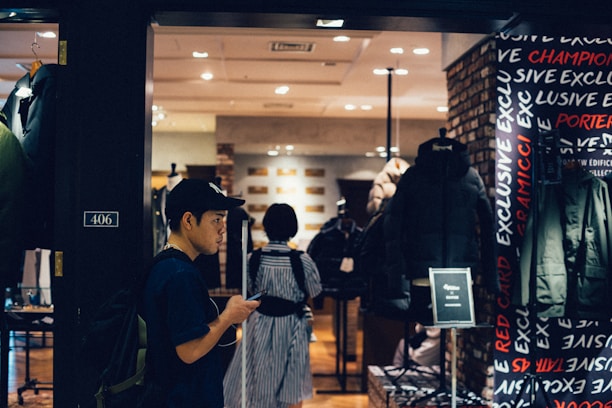 A person wearing a cap and backpack is looking at their phone while walking past a clothing store. The store has a display of jackets and coats, and the interior is well-lit with warm tones. The word 'EXCLUSIVE' is visible on the wall, and there is a black puffer jacket prominently displayed.