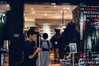 A person wearing a cap and backpack is looking at their phone while walking past a clothing store. The store has a display of jackets and coats, and the interior is well-lit with warm tones. The word 'EXCLUSIVE' is visible on the wall, and there is a black puffer jacket prominently displayed.
