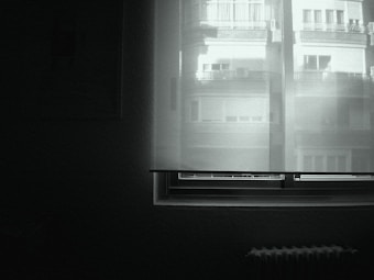 The image displays a minimalist view of a window with closed blinds, through which faint outlines of a building are visible. The room is dimly lit, creating a serene and quiet atmosphere.