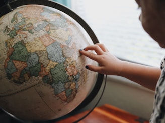 A child's hand with dark nail polish touches a globe, focusing on Africa. The globe is detailed with countries and borders, and it rests on a wooden table beside a window letting in soft light.
