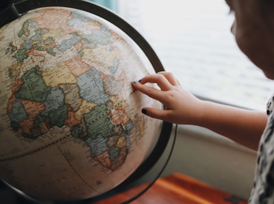 A child's hand with dark nail polish touches a globe, focusing on Africa. The globe is detailed with countries and borders, and it rests on a wooden table beside a window letting in soft light.