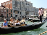 A gondola navigates a canal with a few passengers on board, including a gondolier wearing a striped shirt. The surrounding scenery includes historic Venetian architecture with ornate facades. A larger boat is also visible in the background.