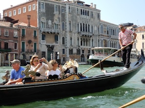 A gondola navigates a canal with a few passengers on board, including a gondolier wearing a striped shirt. The surrounding scenery includes historic Venetian architecture with ornate facades. A larger boat is also visible in the background.